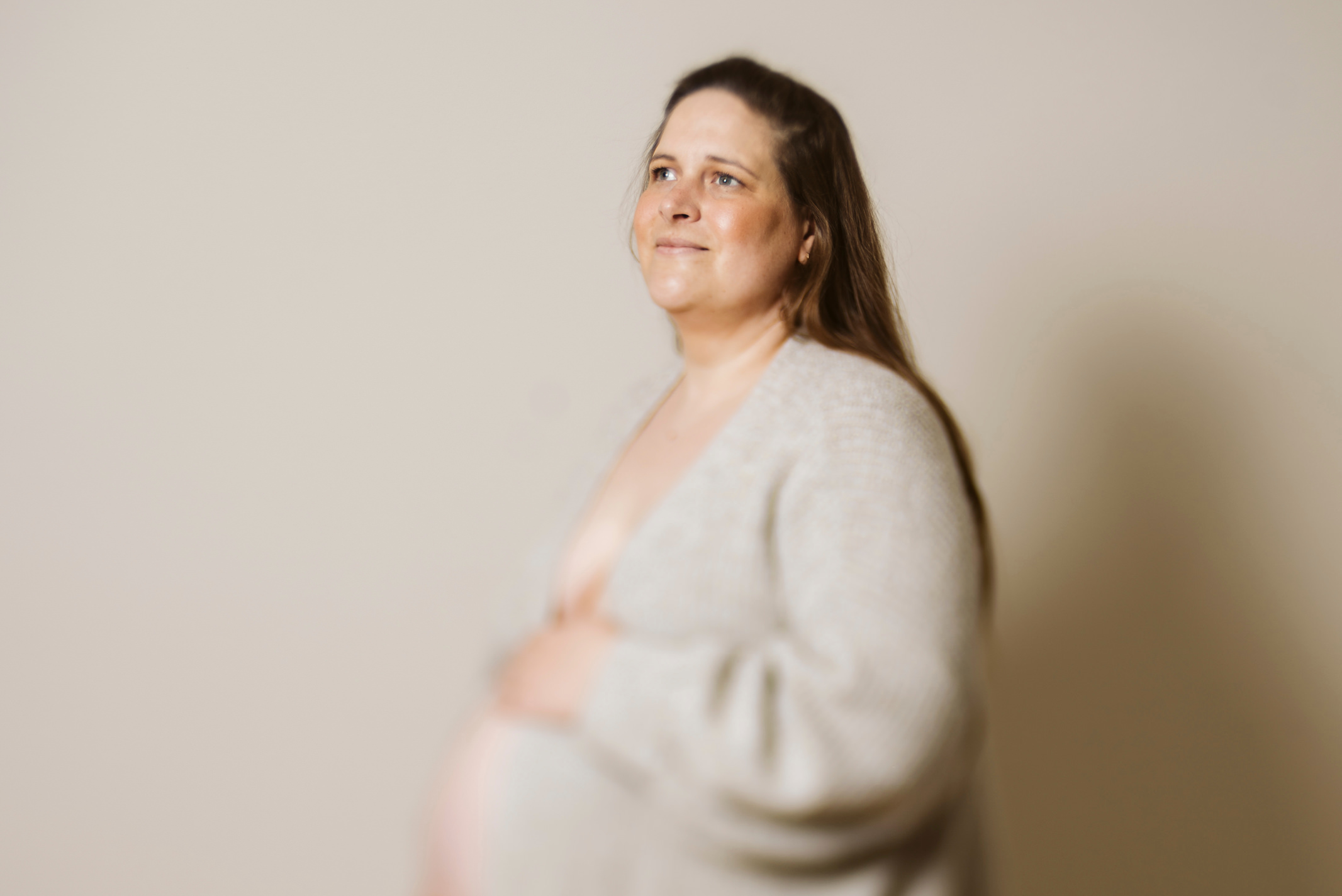 Schwangere Frau in hellem Strick vor beigem Hintergrund. Sie hat eine Hand auf ihrem Babybauch und blickt hoffnungsvoll nach vorn.