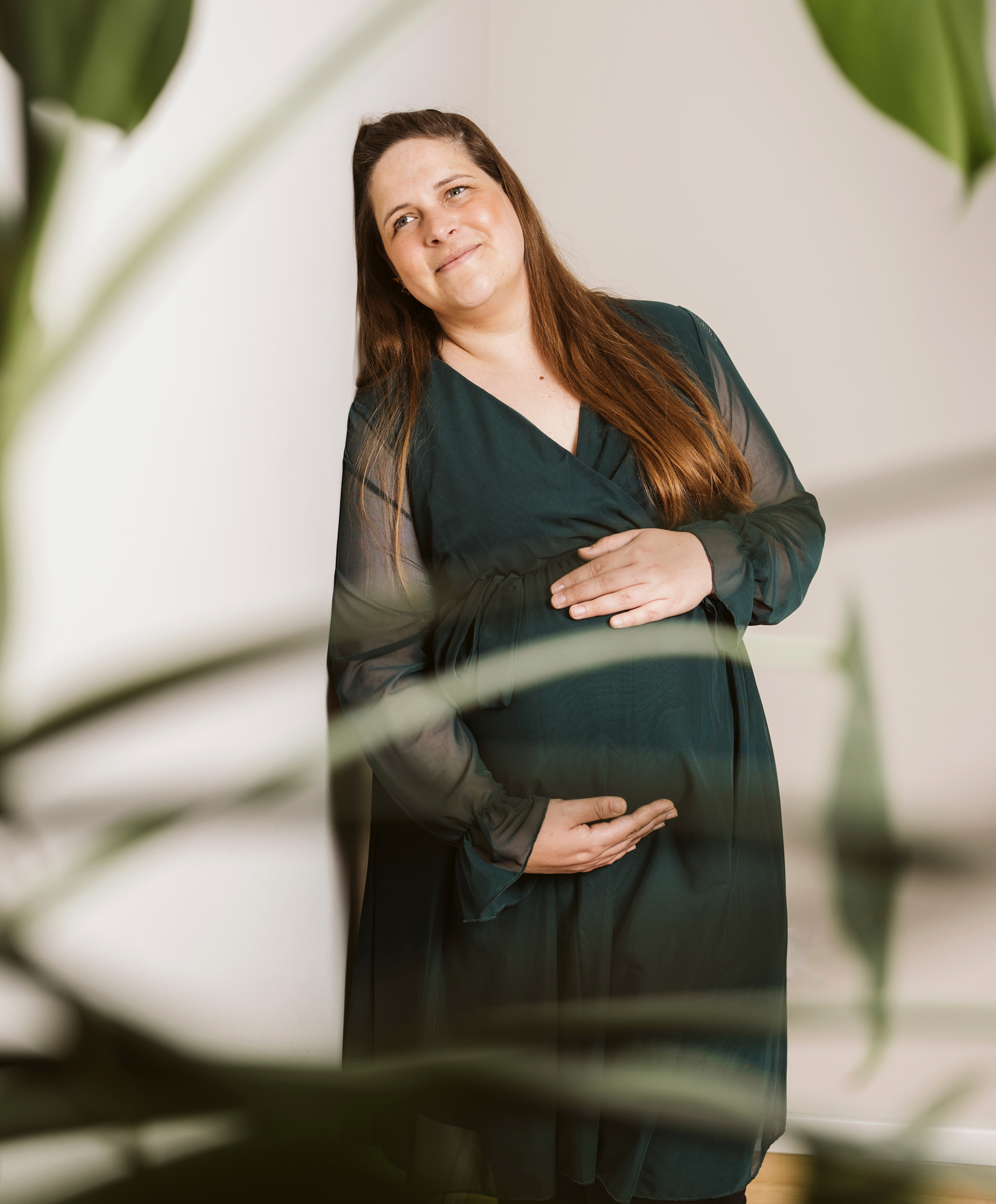 Schwangere Frau in dunkelgrünem Kleid. Sie steht an einer Wand in zartem Braunton. Im Vordergrund sind in Unschärfe Teile einer Monstera-Pflanze zu sehen.