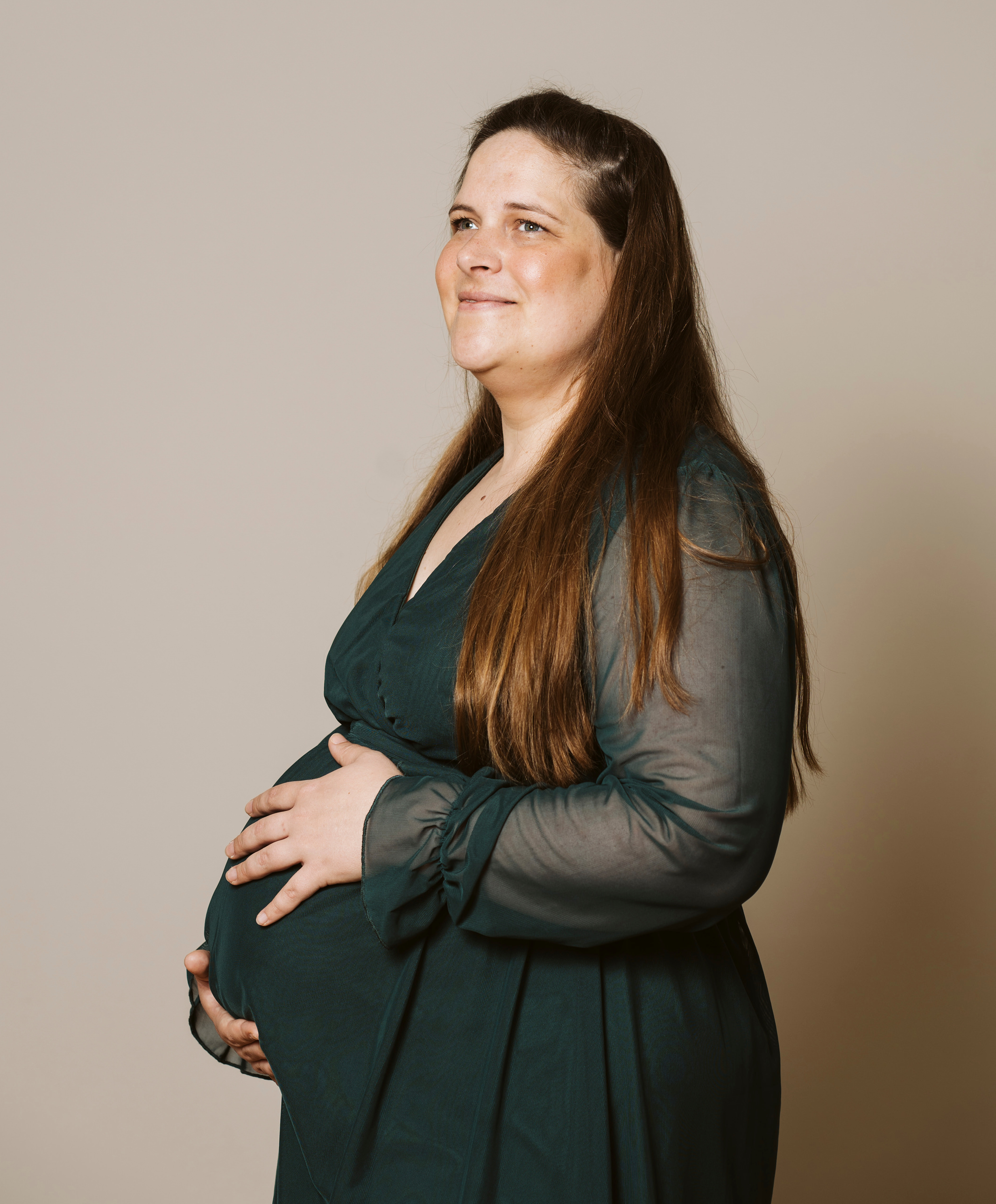 Schwangere Frau in dunkelgrünem Kleid vor einem neutralen beigen Hintergrund. Sie hält ihren Babybauch fest.