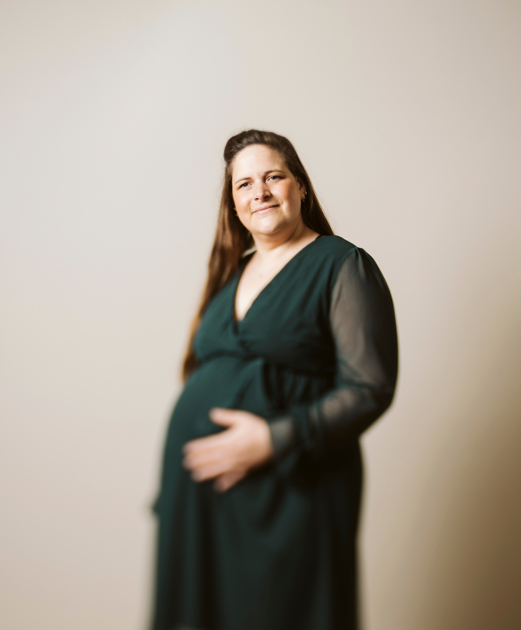 Schwangere Frau in dunkelgrünem Kleid vor einem neutralen beigen Hintergrund. Sie hält ihren Babybauch fest. Teile des Bildes sind bewusst in Unschärfe.
