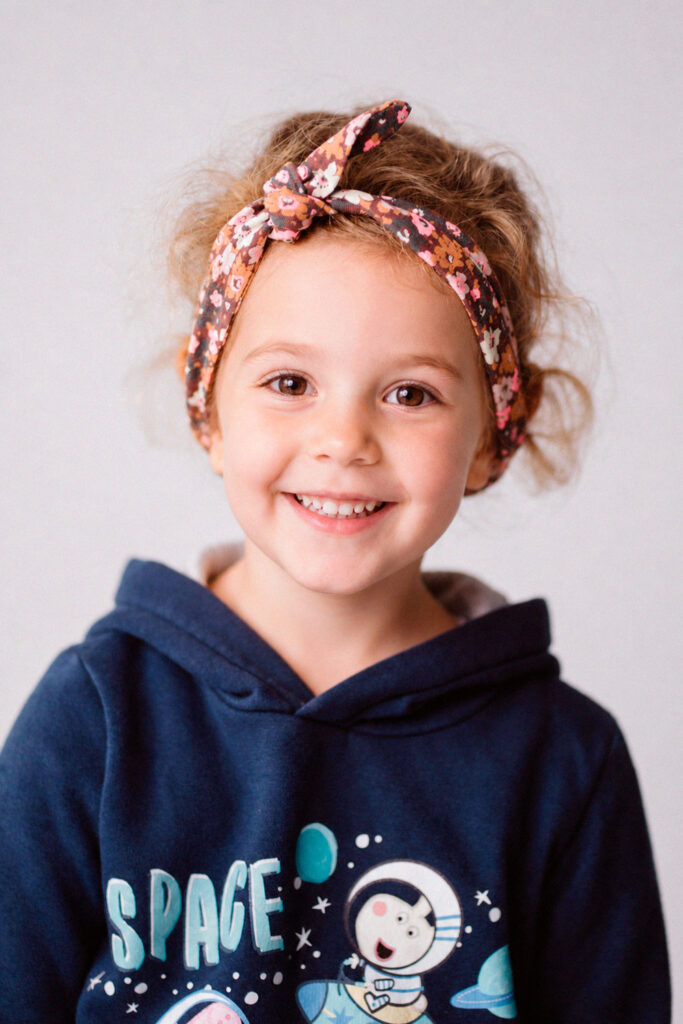 Lächelndes Kindergartenkind. Das Mädchen hat ein Tuch am Haaransatz und einen Hoodie mit Peppa Wutz Aufdruck.